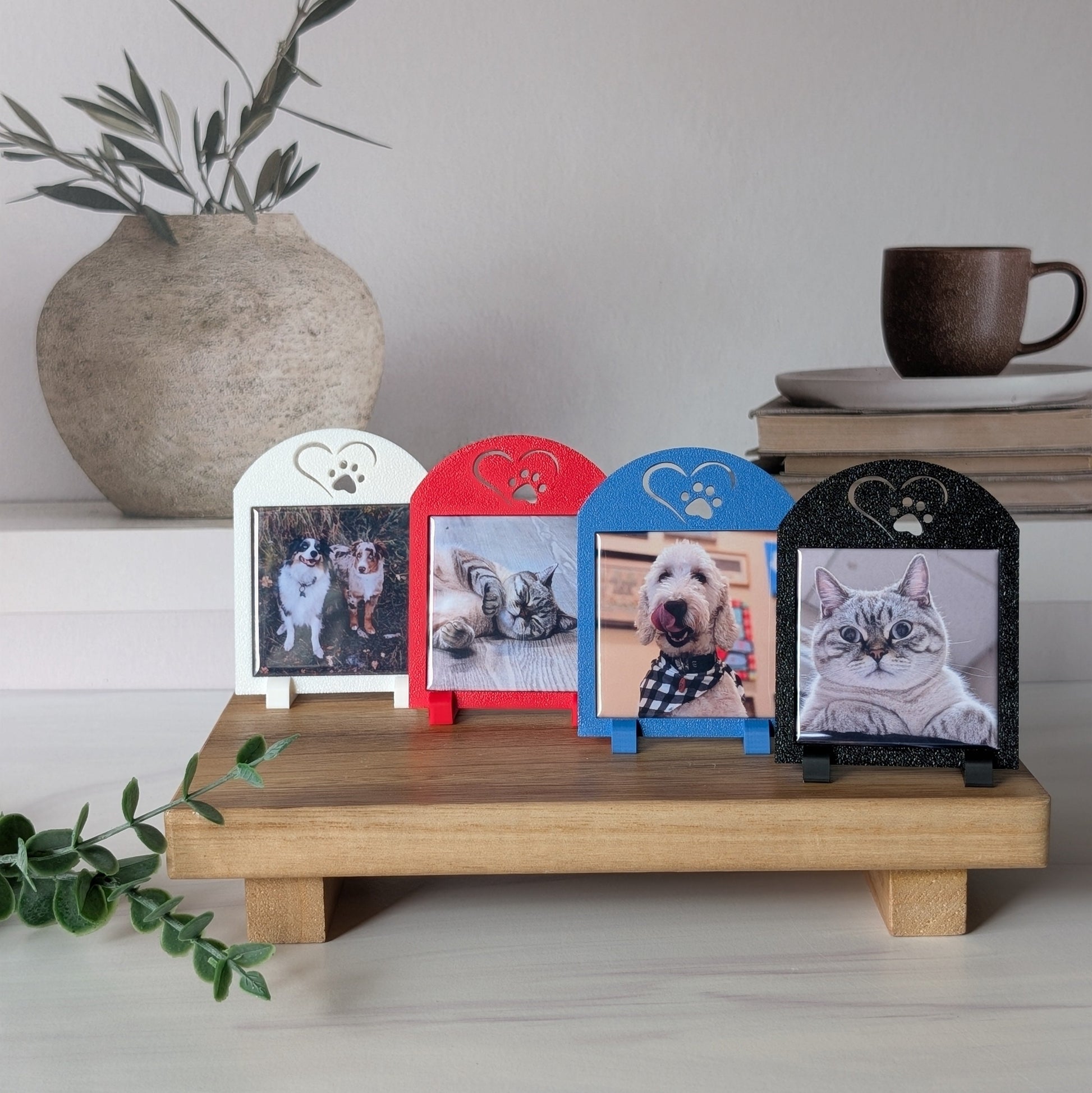 A set of four square photo magnet frames with an icon on the top of a heart outline around a paw. The colors of the frames are white, red, black, and blue. They are sitting on a table.