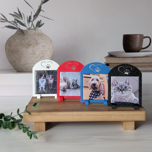 A set of four square photo magnet frames with an icon on the top of a heart outline around a paw. The colors of the frames are white, red, black, and blue. They are sitting on a table.